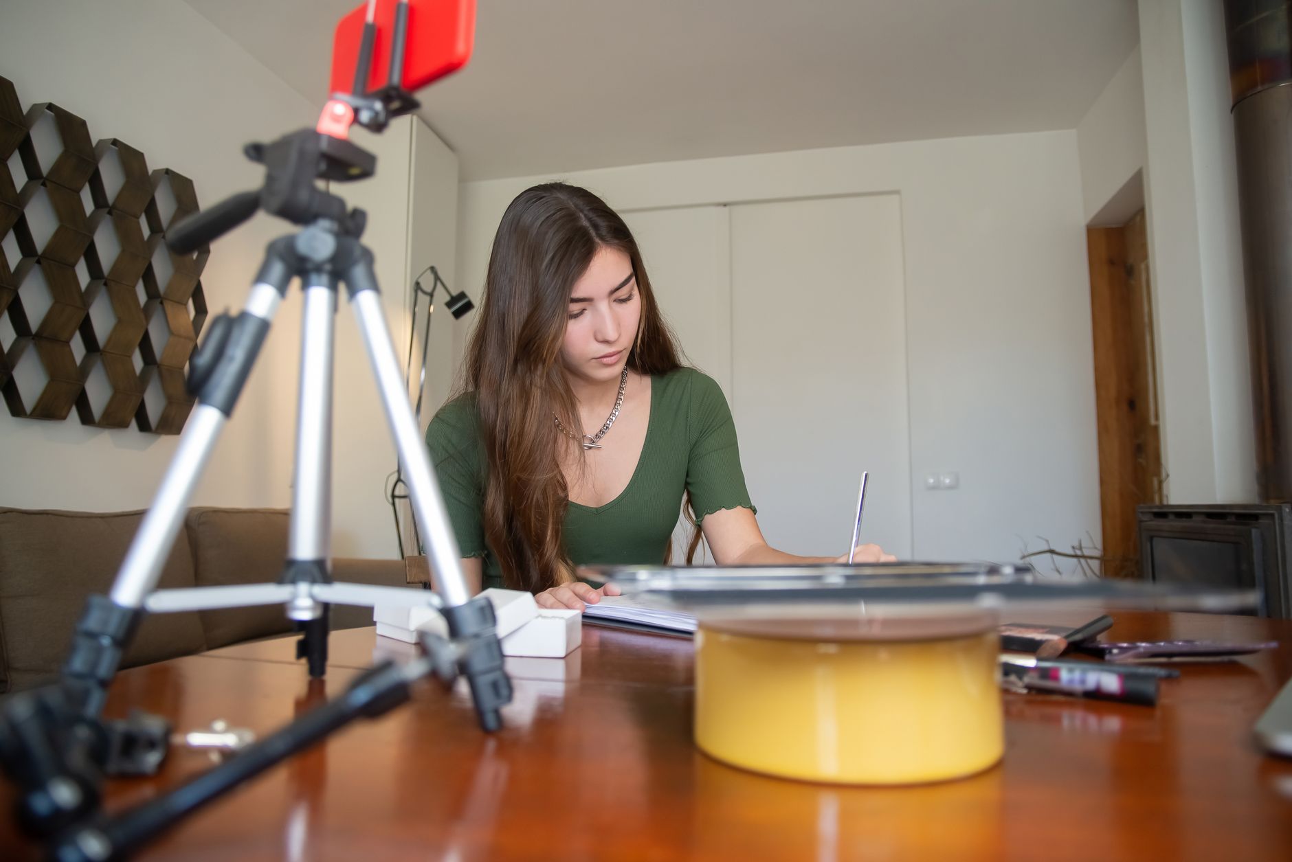 Een jonge vrouw zit thuis aan een houten tafel, met op de voorgrond een smartphone op een statief, klaar om een productvideo op te nemen.
