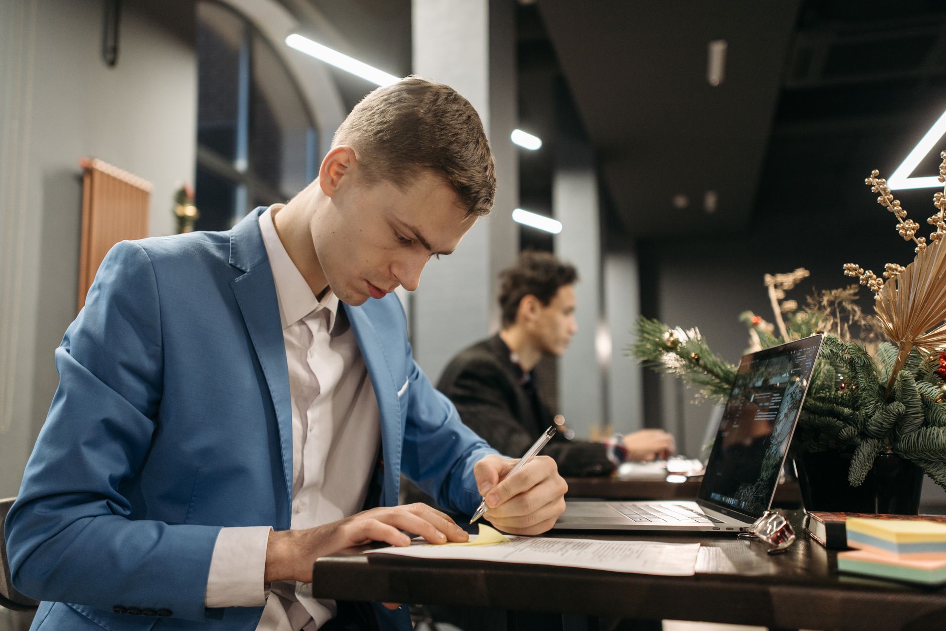 Un homme en veste bleue écrit des notes sur une feuille, à côté d’un ordinateur portable, dans un espace de travail